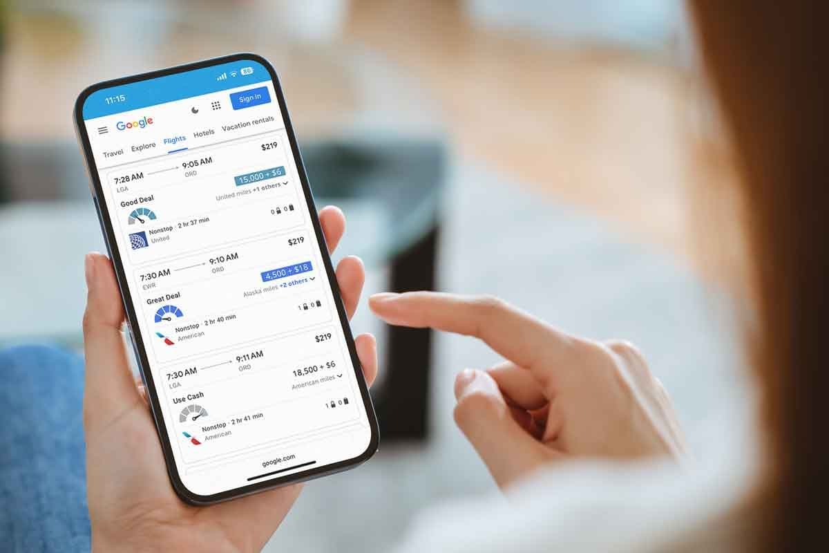 A hand holds an iPhone with Google Flights paired with the Points Path Pro browser extension on the screen, showing award flight prices from New York to Chicago along with a meter for whether or not they're a good deal.