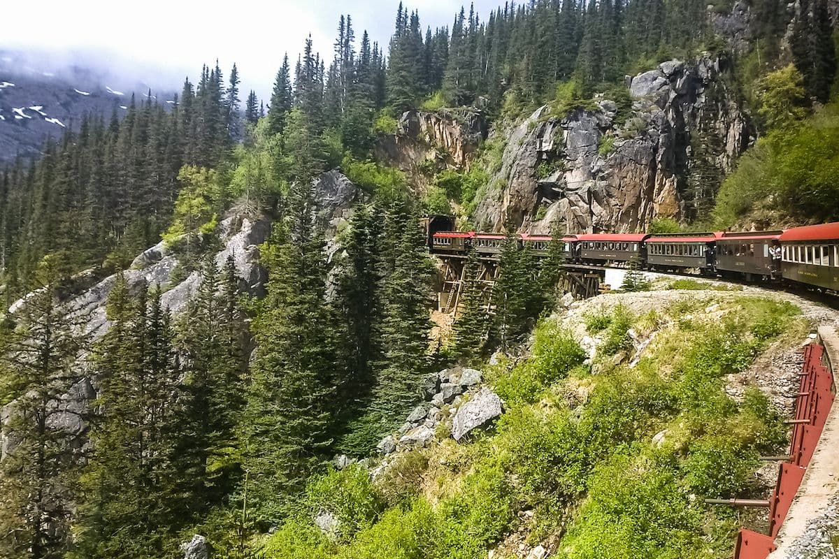 Train cars crossing a narrow trestle along a steep, forested mountainside on the White Pass Scenic Railway near Skagway, Alaska, surrounded by evergreen trees and rocky cliffs.
