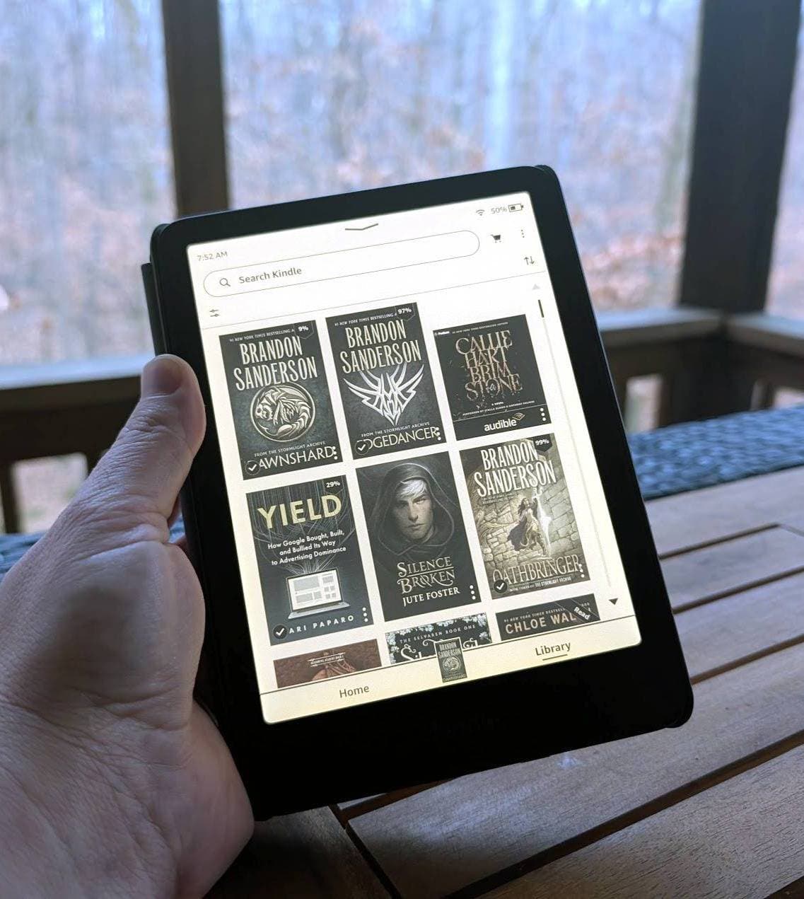 Person holding a Kindle e-reader displaying a digital book library while sitting outdoors on a porch.