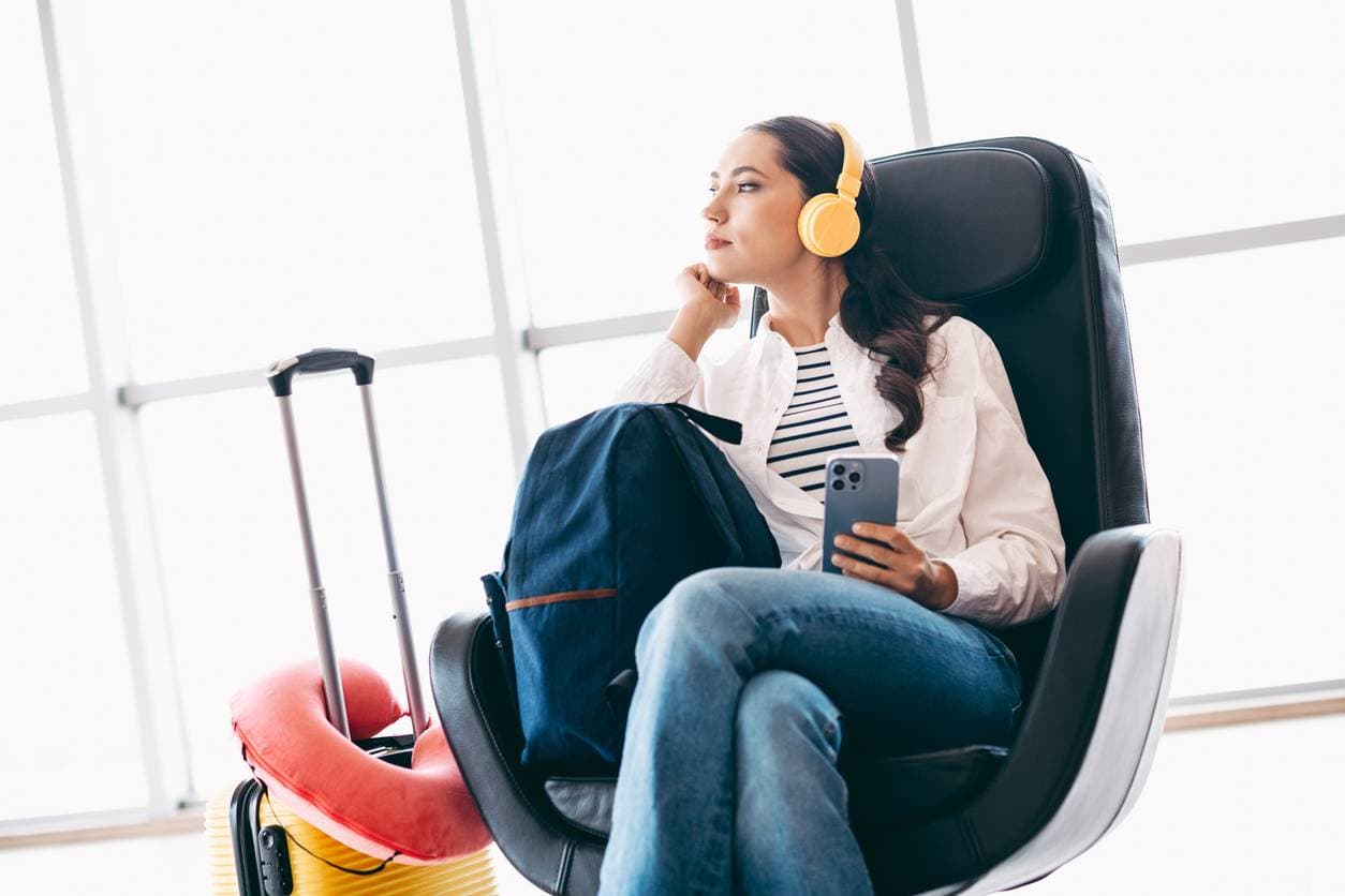 A woman wearing yellow headphones sits in a leather chair near an airport window, holding her phone with a yellow suitcase and travel pillow beside her.